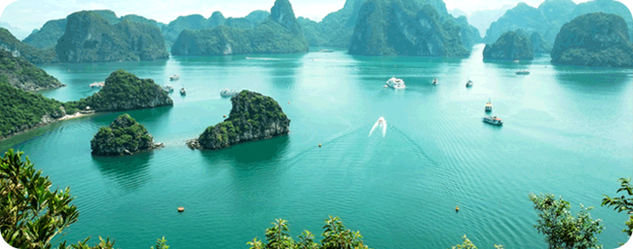 Halong Bay. A beautiful blue lagoon with a group of islands in the background. The water is calm and clear, with several boats floating on the surface. The sky is blue and cloudless, creating a perfect day for boating and relaxing.